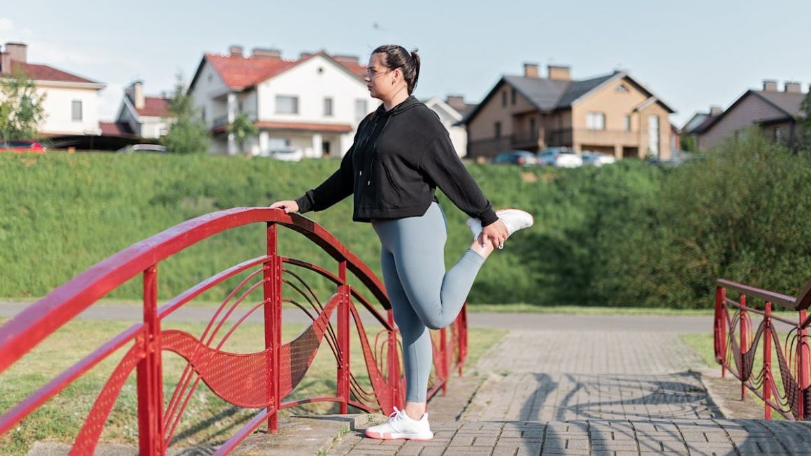 A woman in active wear performs a stretching exercise on a red bridge in a suburban park setting.